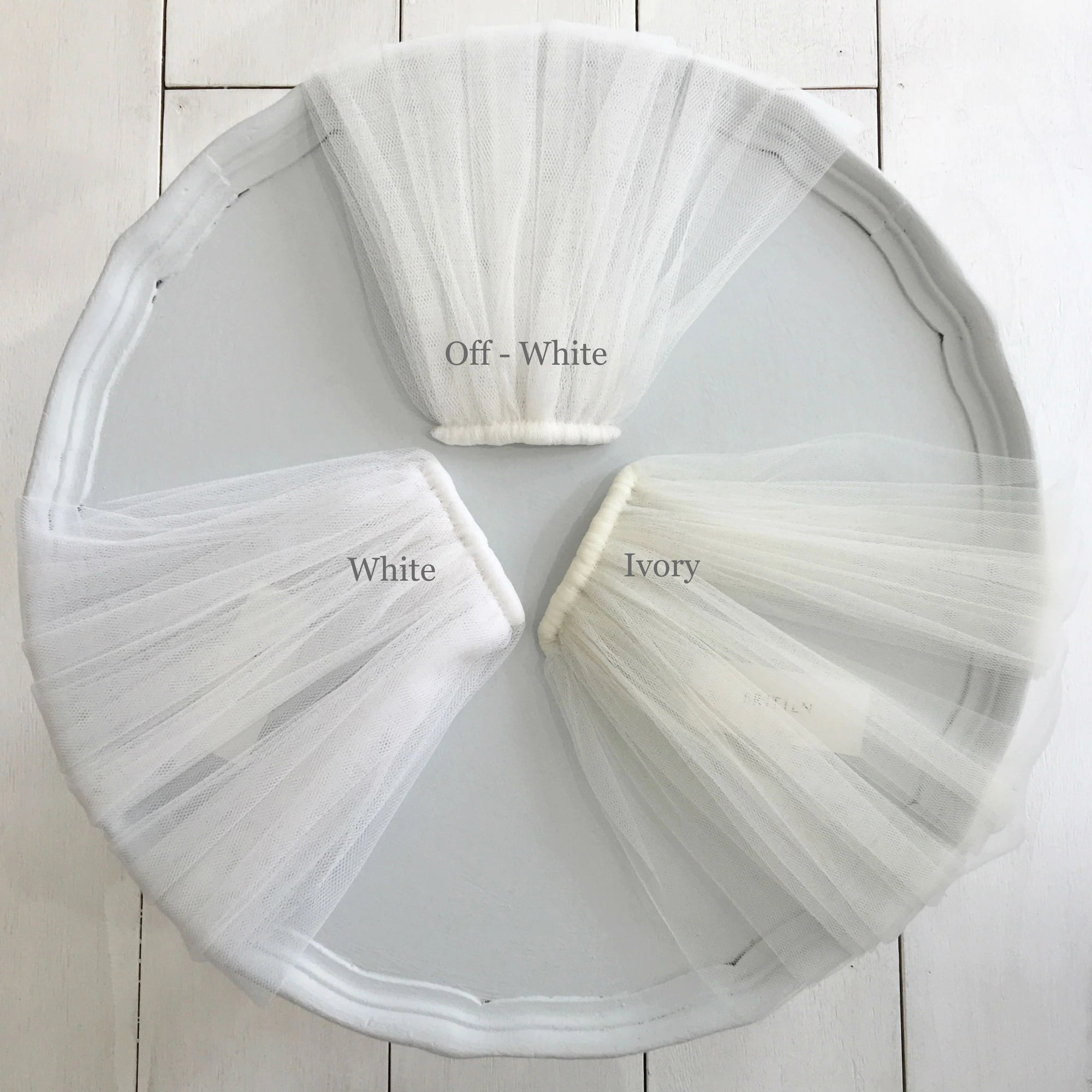 Wedding veil tulle fabric color comparison – three gathered tulle samples labeled White, Off-White, and Ivory, displayed on a round tray. A guide to choosing the perfect veil shade to match a wedding dress.